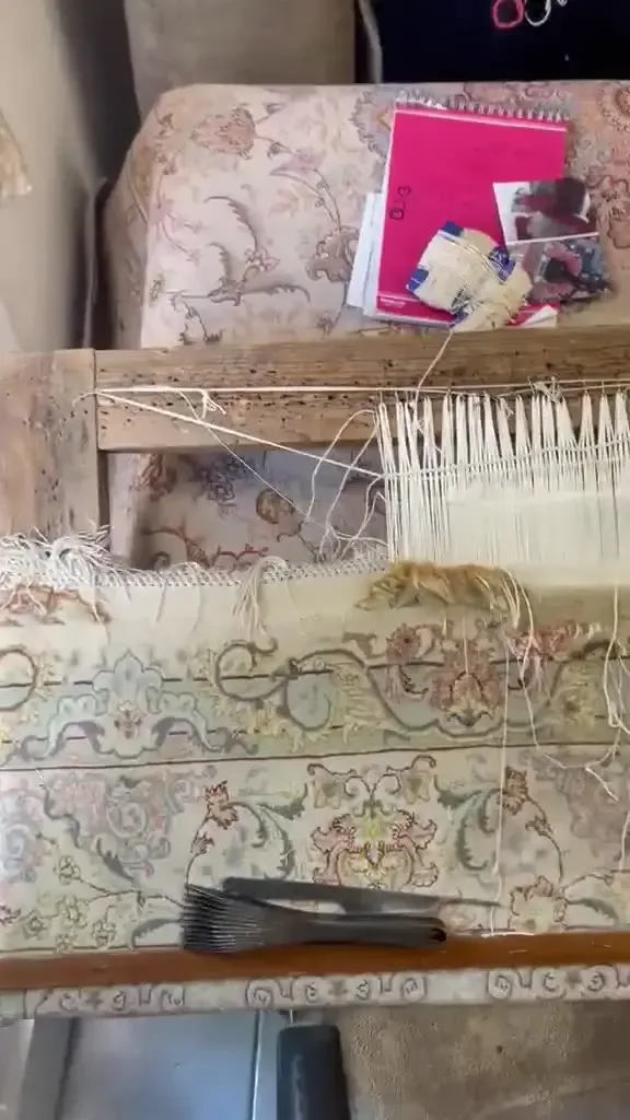 Bord d'un tapis clair à motifs floraux, fils blancs tendus sur une barre en bois, carnet rose et outil à dents métalliques posés dessus