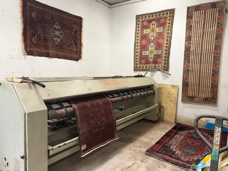 Machine à rouleaux avec un tapis bordeaux engagé à l'avant, trois tapis accrochés au mur blanc derrière et un tapis posé au sol à droite